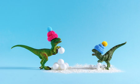 Cute green dinosaur in knitted hats playing snowballs. Happy winter holidays card.の写真素材