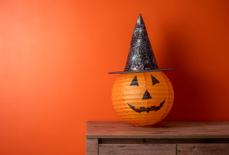 Paper lampshade with spooky face in witch hat on wooden table against orange wall. Space for text. Halloween minimal greeting card.の写真素材