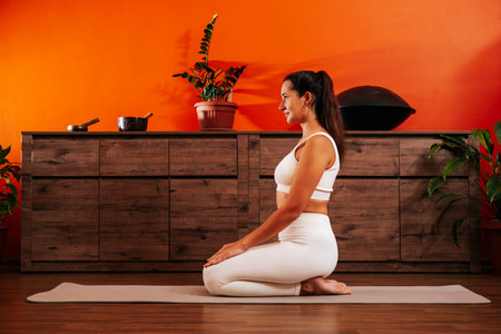 Young beautiful woman with straight back and good posture sits on yoga mat sideways to the camera against the background of a wooden chest of drawers and an orange wall.の写真素材