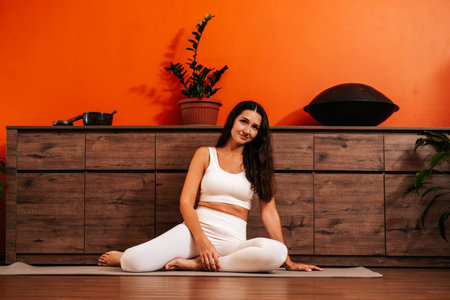 Beautiful young woman in white sports clothes is sitting on yoga mat against background of wooden furniture and orange wall in yoga class. Female wellbeing concept.の写真素材