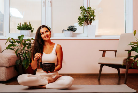 Happy beautiful young woman is sitting in lotus position and palyying singing bowl on yoga mat at yoga class. Soundhealing concept.の写真素材