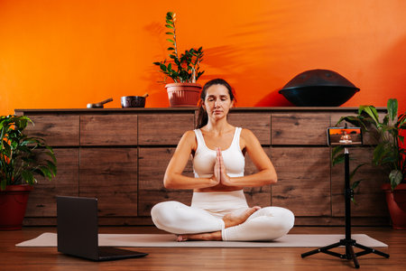 Beautiful young brunette woman is sitting in lotus position with her hands in namaste on yoga mat at home in living room.の写真素材