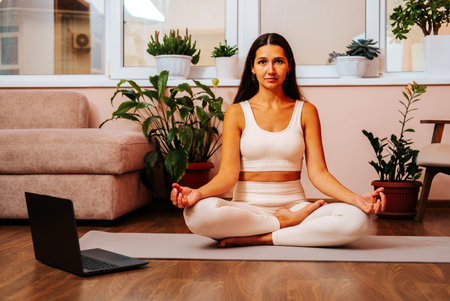 Beautiful young brunette woman is sitting in lotus position on yoga mat at home in living room.の写真素材