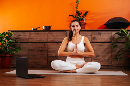 Young woman sitting in lotus position with hands in namaste and doing yoga online.の写真素材