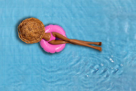 Top view doll in straw hat and pink bikini floating on inflatable circle in blue water of swimming...