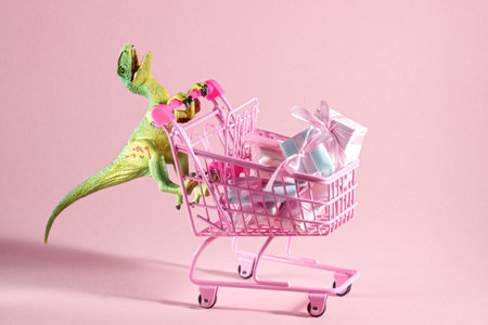 Funny green dinosaur toy with shopping cart full of present boxes on a soft pink background.の写真素材