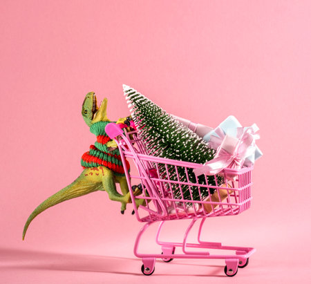Happy green dinosaur carrying shopping cart with Christmas tree and gift boxes on pink background.の写真素材