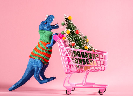 Cute blue dinosaur carrying shopping cart with Christmas tree on pink background.の写真素材