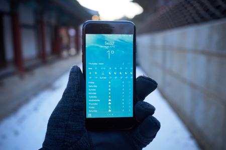 Seoul, South Korea - December 13: A gloved hand is holding a black smartphone. The smartphone is showing a weather forecast for Seoul. The weather forecast is showing 1 degrees celcious. The background is out of focus and blur.のeditorial素材
