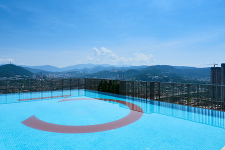 A swimming pool located on a tall building. The swimming pool is located at the edge of the building. The background consists of nature landscape, mountain, and some low density apartment buildings.の写真素材