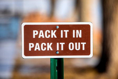 a brown, pack it in, pack it out sign with white lettering and white boarder. Taken at Cheery Creek reservoir in Colorado. With snow trees in the background.の写真素材