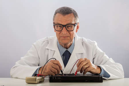An older male wearing a white lab coat and repairing electronic equipments, like a technician or a...