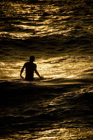 An image of a young man on Maui, standing in the Pacific Ocean, bathed by the golden light of the setting sun.の写真素材