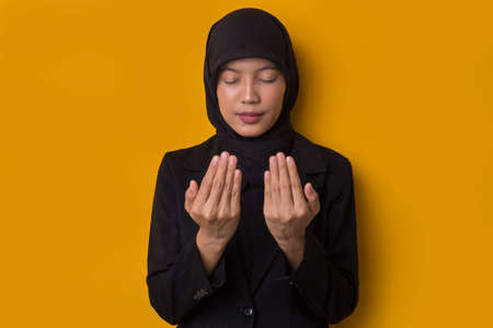 Young asian muslim woman with hijab praying on yellow background