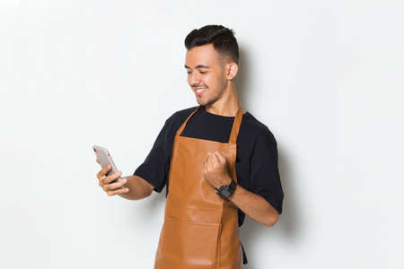 Happy joyful young man barista bartender or waitress using mobile smartphone isolated on white backgroundの写真素材