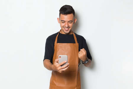 Happy joyful young man barista bartender or waitress using mobile smartphone isolated on white backgroundの写真素材