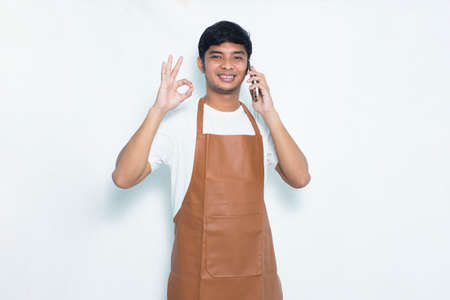 Happy joyful young asian barista man bartender or waitress using mobile smartphone isolated on white backgroundの写真素材