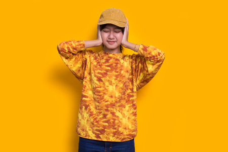 young asian beautiful woman covering both ears with hands isolated on yellow backgroundの写真素材