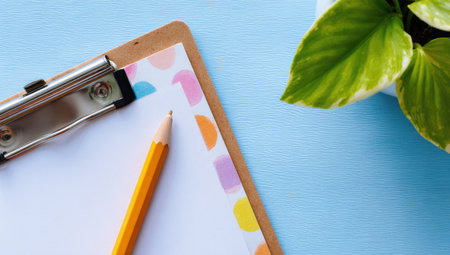 Clipboard with a sheet of paper featuring a colorful design of circles and a pencil resting on it.  A small plant is visible in the top right corner.の素材