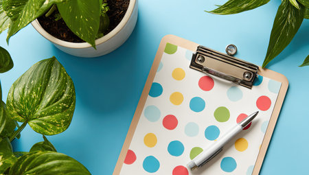 Clipboard with a colorful polka dot pattern rests on a light blue surface.  A white pen lies beside it.  A potted houseplant with large leaves adds a touch of nature to the image.の素材