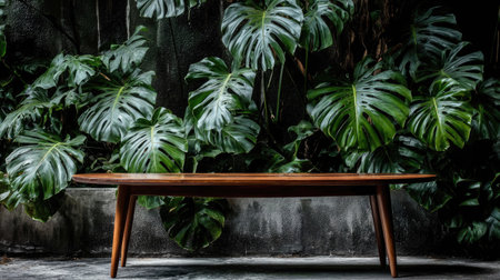 Long, oval, wooden coffee table, with simple, clean lines and a warm brown hue, is placed in front of a wall of lush, vibrant green tropical foliage.  The concrete floor and wall are a muted gray. The image has a calm, serene aesthetic.の素材