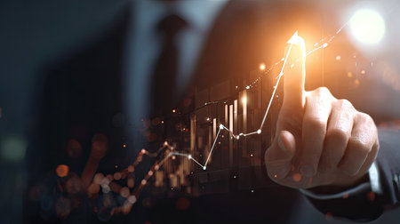 View of a person in formal business attire, presumably an executive or manager, using a finger to interact with a holographic or digital display showing a financial line graph with an upward trajectory and accompanying bar charts, set against a dark, bokeh-filled background with warm orange lighting emphasizing growth and success.の素材