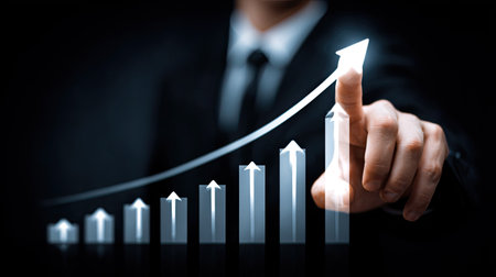 Shot focuses on a business person in a dark suit and tie, whose hand is interacting with a holographic or digital bar graph. The bars show a clear progression in height, culminating in a bright, upward-pointing arrow that the index finger appears to be touching, symbolizing positive momentum, achievement, and future success in an economic or corporate context against a deep, dark backdrop.の素材