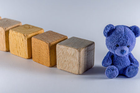 Composition featuring a row of wooden blocks of various shades of brown and a small, sitting blue teddy bear against a white background.  The blocks are arranged in a line, while the teddy bear sits to the right of the blocks. The image is well-lit and presents a clean, simple aesthetic.の素材