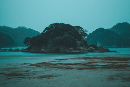A small, tree-covered island sits in calm, blue water with misty hills behind.の素材
