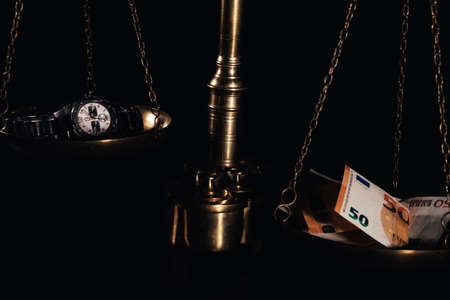 a golden scale on a black background, on one side there is a clock representing time, on the other side there are banknotes representing money, concept the value of time and money, in the image the money with more weight.の写真素材