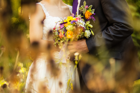 The groom and the bride are holding a bouquet in the park among the grassの写真素材