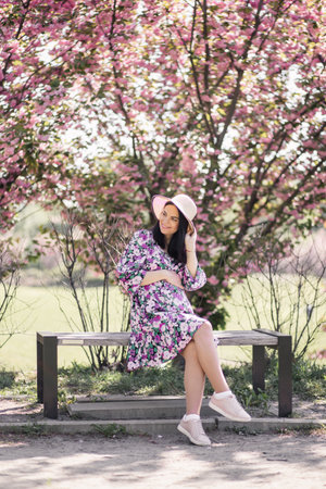 pregnant girl sits in a dress on a bench near cherry blossomsの写真素材