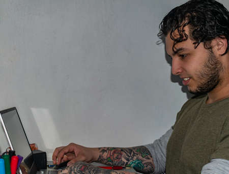 white man with black curly hair, working at home from laptop, smiling, with tattoos on both arms and beardの写真素材