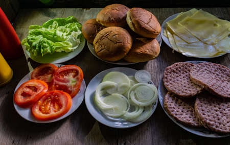 Ingredients for preparing hamburger, on white plates, bread, onion, red tomato, raw meat, cheese and lettuce, with tomato sauce and mustard on the left side on wooden boardの写真素材