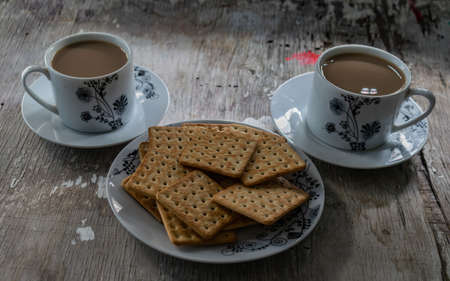 square cookies on white plate, with two cups of coffee with milk in the backgroundの写真素材