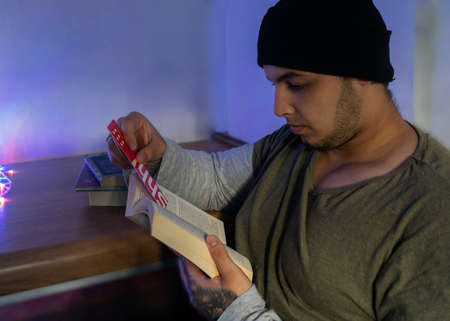 Young white man with a beard, winter hat and sweater, sitting reading a book, leaning on a table, with three books on it and colored lightsの写真素材