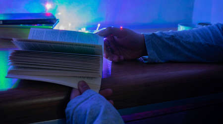 Hands of a white man, holding a book, with three books in the background and colored lights, on a wooden table, with a white backgroundの写真素材