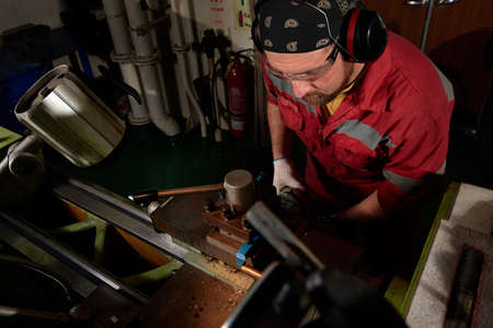 Worker in red uniform operating in manual lathe in metal big workshop with dark environment.