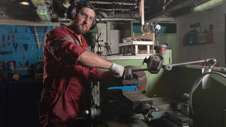 Worker in red uniform operating in manual lathe in metal big workshop with dark environment.