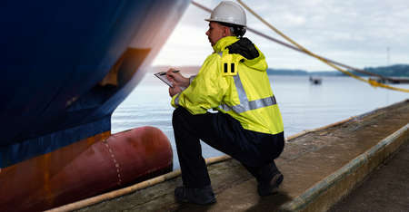 Ship supervisor engineer inspector stands at the dockside in a port. Wearing safety helmet and yellow vest. Cargo shipping industry. Protection and idemnity concept.の写真素材