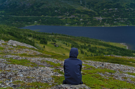 Back view boy child sitting alone on gray rock, looks down the mountain to the green valley and blue lake. Northern Europe Gjevilvatnet Norwayの写真素材