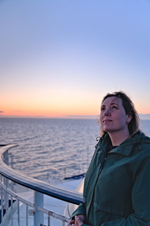 Beautiful woman leaning against the railing of the boat, looking at the sky with a satisfied face with the wind in her hair. Glowing sunset on the sea summer vacation on the Scandiの写真素材