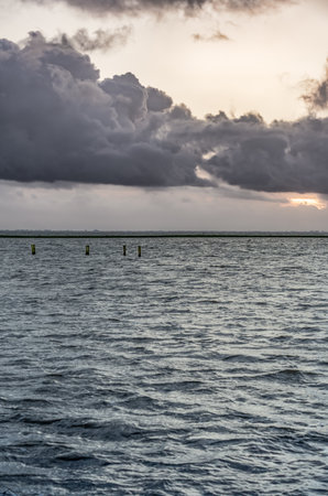 Tranquil waters reflect the soft light of dusk with wooden poles emerging from the flooded overflow area. Dike in the distance separates the calm lake from the dramatic evening skyの写真素材