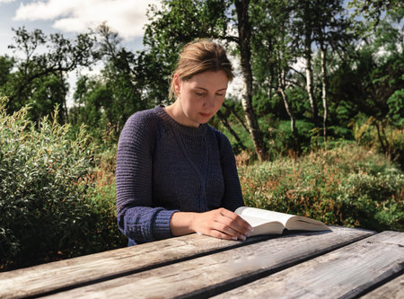 A woman sits at a wooden table, deeply engrossed in reading a book surrounded by vibrant greenery and trees, enjoying a tranquil moment in the nature of Sweden.の写真素材