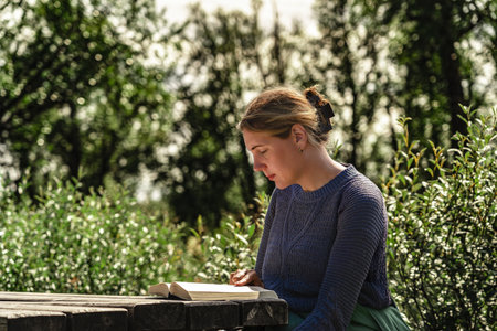 A woman sits quietly at a wooden table, engrossed in a book while surrounded by greenery in a peaceful outdoor setting, enjoying reading in a serene moment in the summer natureの写真素材