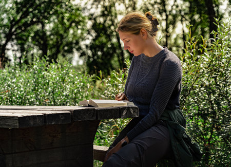 Beautiful woman sits quietly at a wooden table, engaged in a book while surrounded by greenery in a peaceful outdoor setting, enjoying reading in a serene moment in summer natureの写真素材