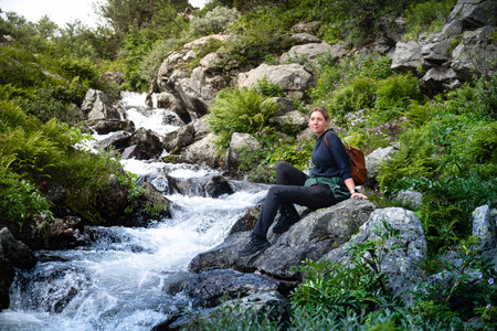 Female hiker taking a break, sitting on a rock beside the rushing stream Silverfallet in Dalarna Sweden, vibrant greenery and rocky terrain, enjoying a peaceful moment in nature.の写真素材