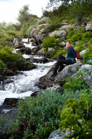 Woman taking a break and relaxing on a rock beside a mountain stream, looking over her shoulder at the vibrant greenery and wildflowers in the natural beauty of nordic wildernessの写真素材