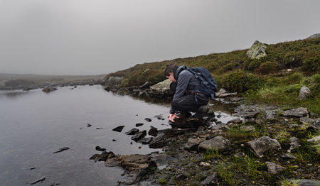 A boy crouches by a mountain lake in Sweden, surrounded by fog and rain. He examines the water's edge while wearing a backpack and enjoying nature high in the mountains.の写真素材