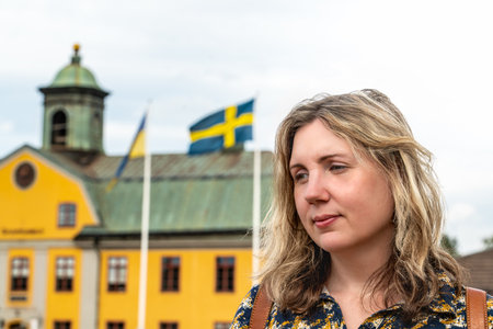 A woman poses thoughtfully with the Swedish and Ukrainian flags in view, set against a colorful building in Sweden during a sunny summer day.の写真素材